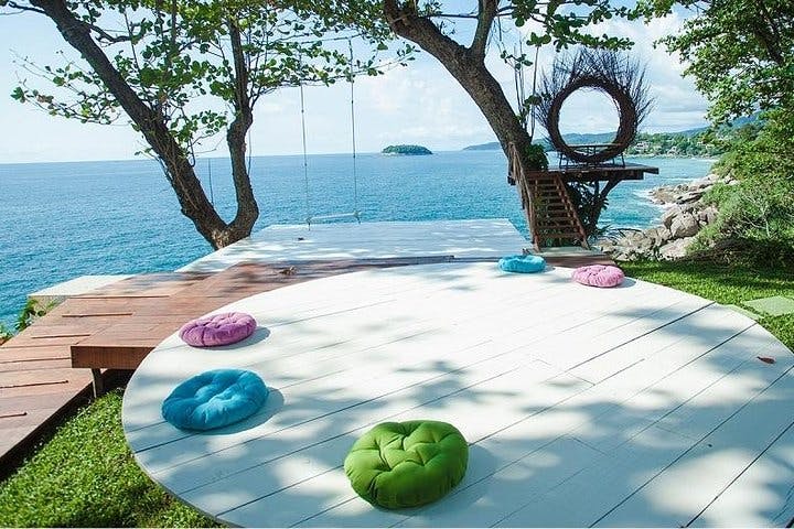 Wooden deck by the sea with colorful cushions, trees with swings, and a raised platform with a nest-like structure.