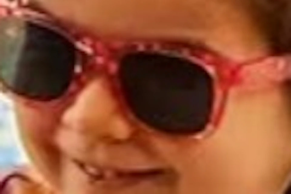 A young girl wearing sunglasses holds a colorful parrot on her arm, with another parrot held by someone nearby.