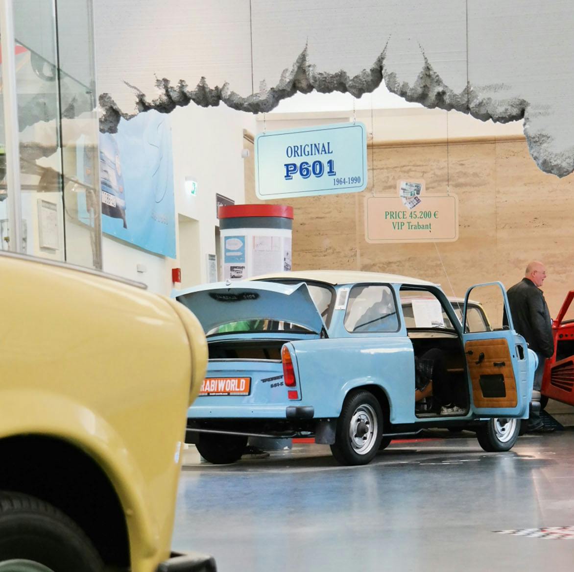A light blue car, with open doors and trunk, is displayed in a showroom. Signs on the wall indicate it is an "Original P601."