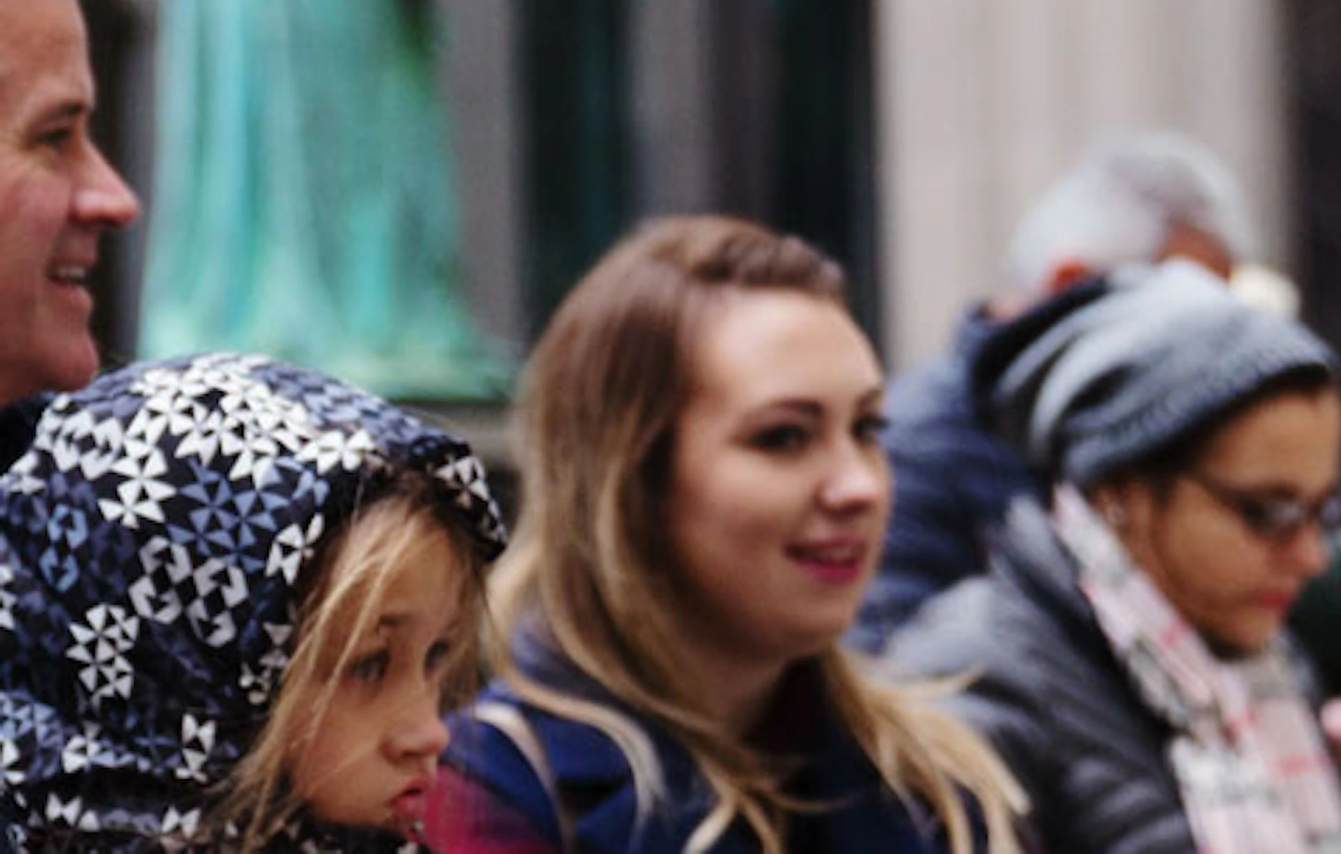 A woman in a red coat and purple headband gestures while speaking to a group of people outdoors in a city setting.
