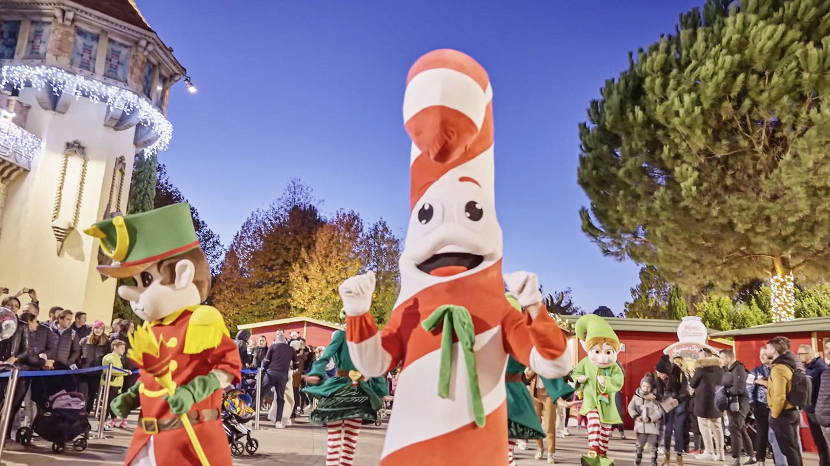 황혼 하늘 아래 배경의 사람들과 함께 축제 야외 행사에 참여하는 지팡이 사탕과 엘프 의상을 입은 캐릭터.