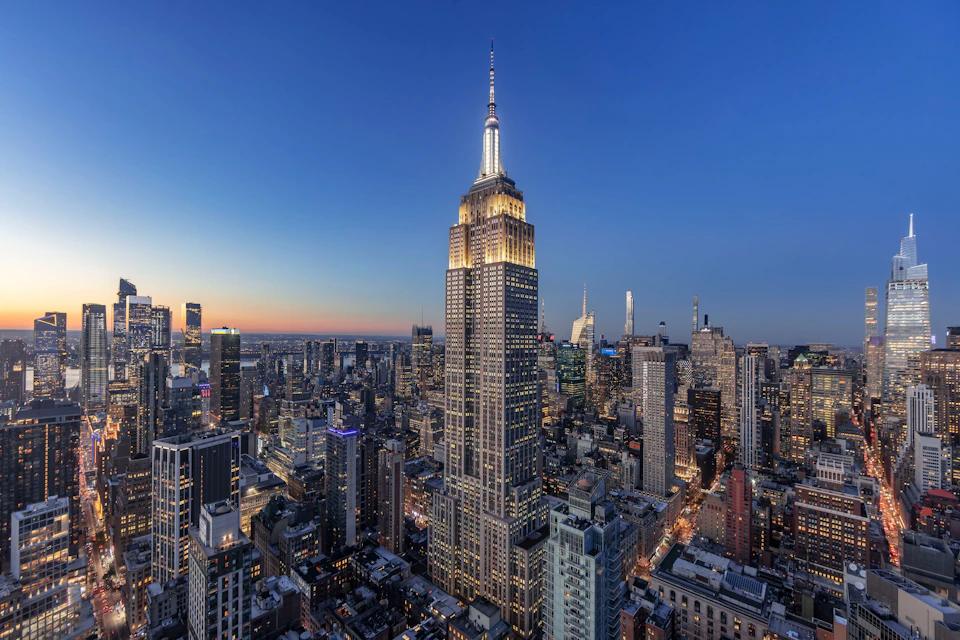 A cityscape at dusk featuring a tall, illuminated skyscraper surrounded by buildings under a clear, dimming sky.