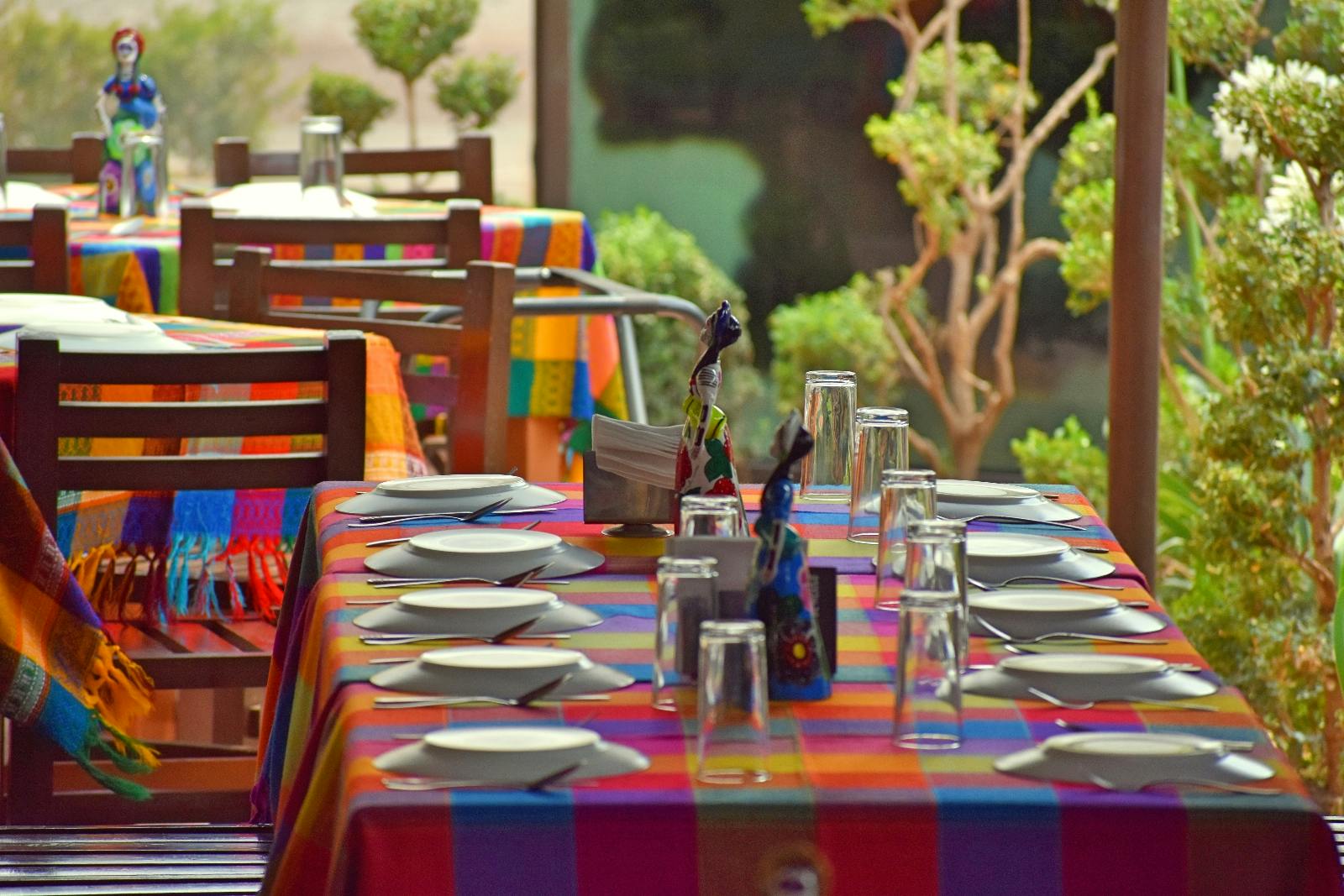 Venkovní jídelní stoly s pestrobarevnými ubrusy, talíři, sklenicemi a dekorativními figurkami, obklopené zelení.