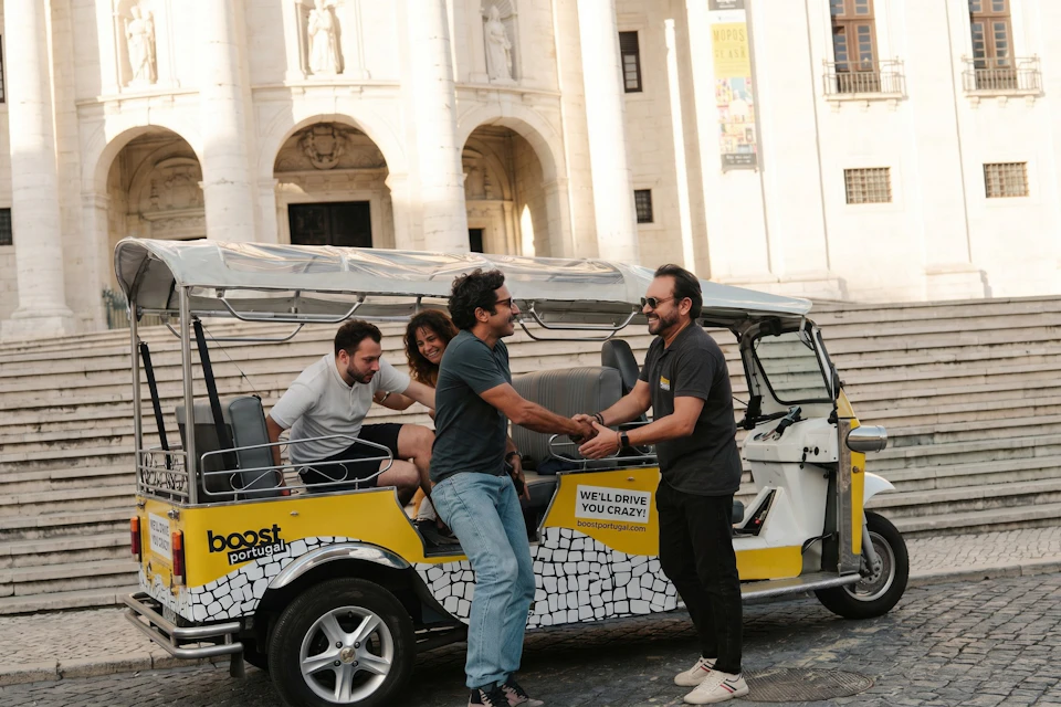 Two people shake hands beside a tuk-tuk styled vehicle with passengers, parked in front of a historic building.