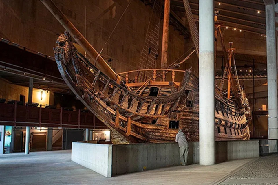 A large wooden ship displayed in an indoor museum setting with a person standing nearby for scale.