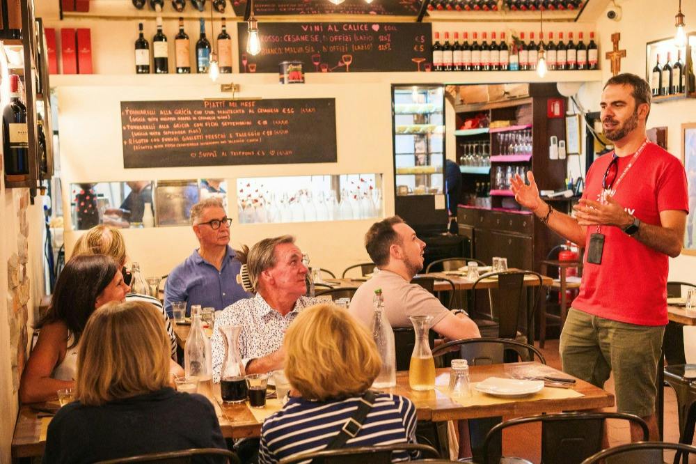 A man speaks to a group of people seated at tables in a cozy restaurant with shelves of bottles and a chalkboard menu.