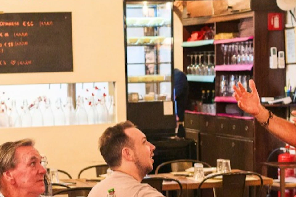 A man speaks to a group of people seated at tables in a cozy restaurant with shelves of bottles and a chalkboard menu.