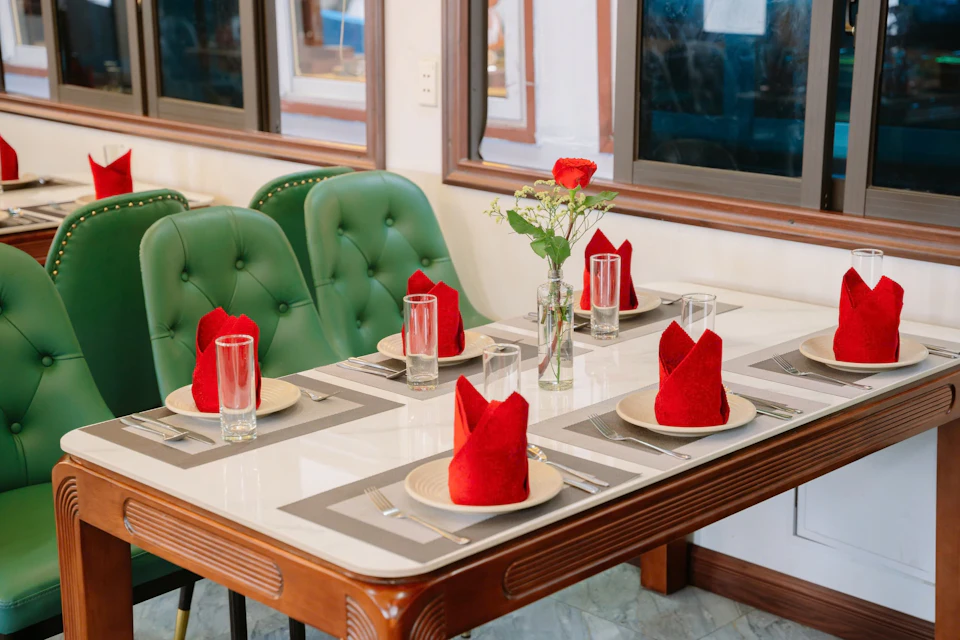 A dining table set for six with red napkins, clear drinking glasses, and a vase with a single red rose and white flowers.