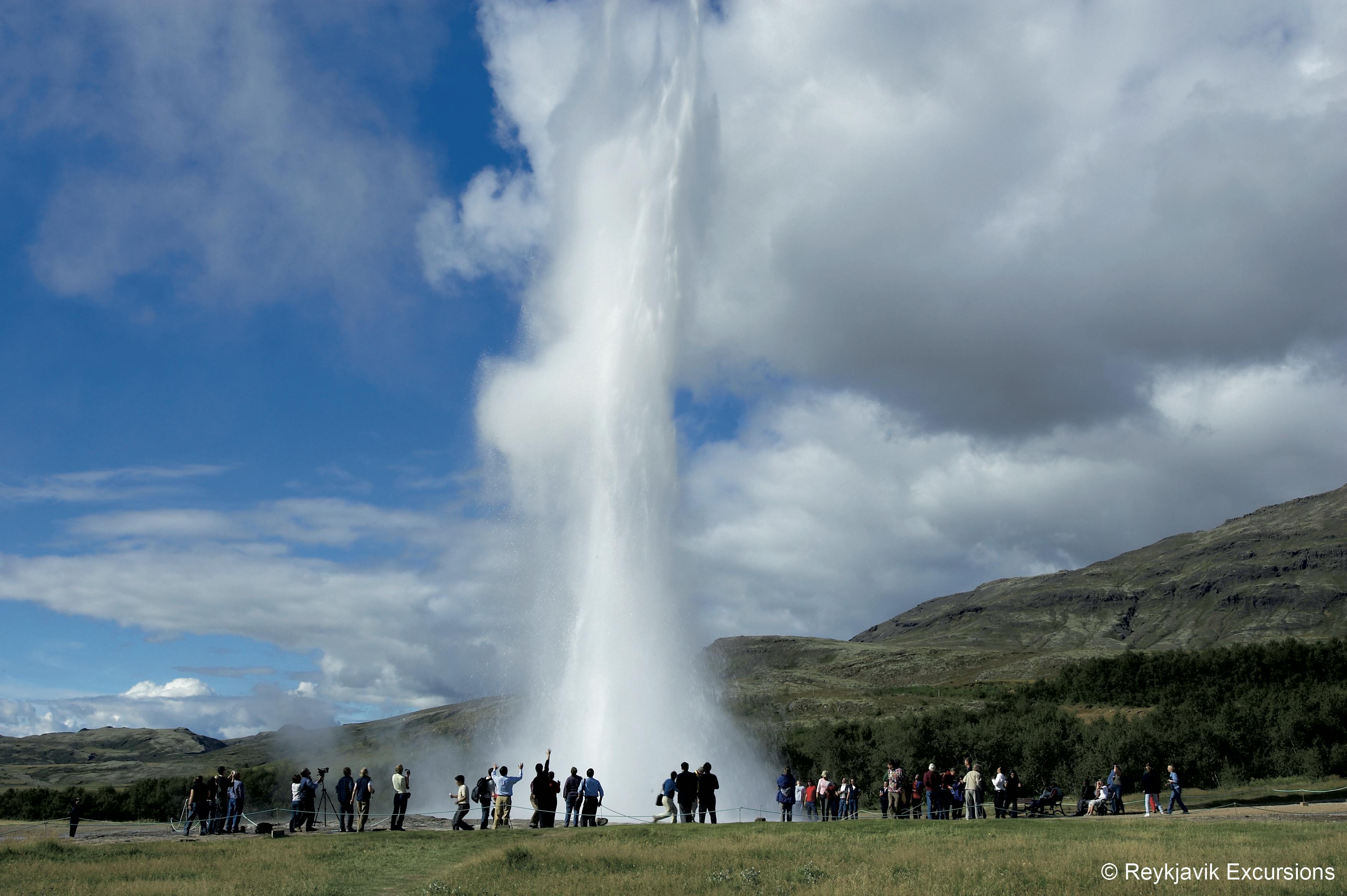 Lidé se shromáždili kolem gejzíru vybuchujícího vysoko do nebe na pozadí travnatých kopců a částečně zatažené oblohy.