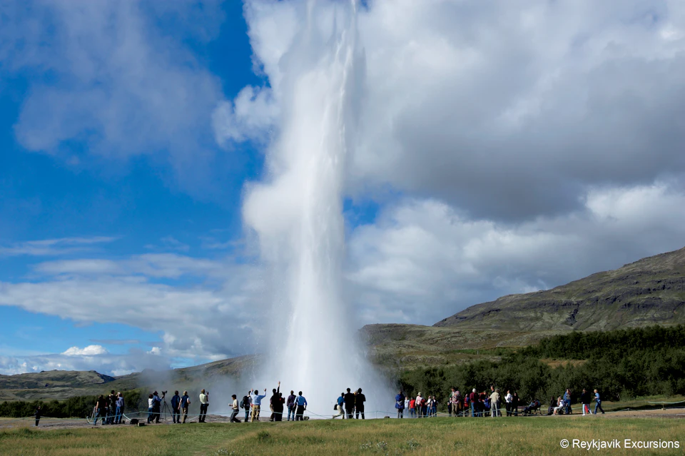 People gathered around a geyser erupting high into the sky against a backdrop of grassy hills and a partly cloudy sky.