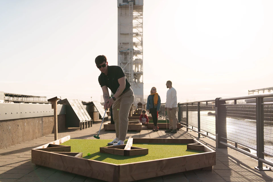 A person plays mini-golf on a rooftop course with others nearby and a tower structure in the background under bright sunlight.
