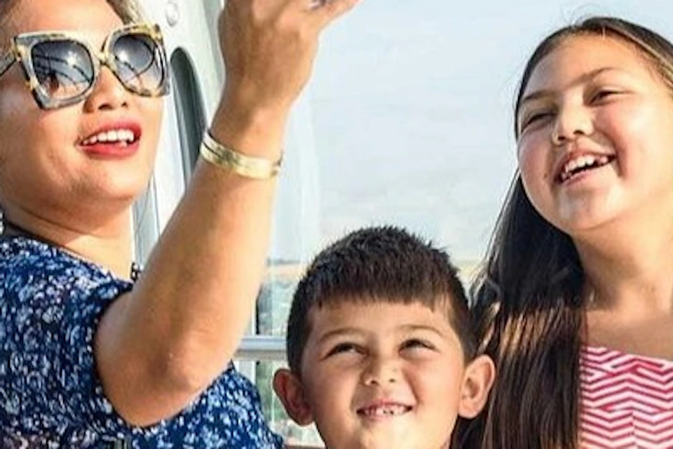 A woman and two children smiling and taking a selfie inside a glass observation capsule with a city view in the background.