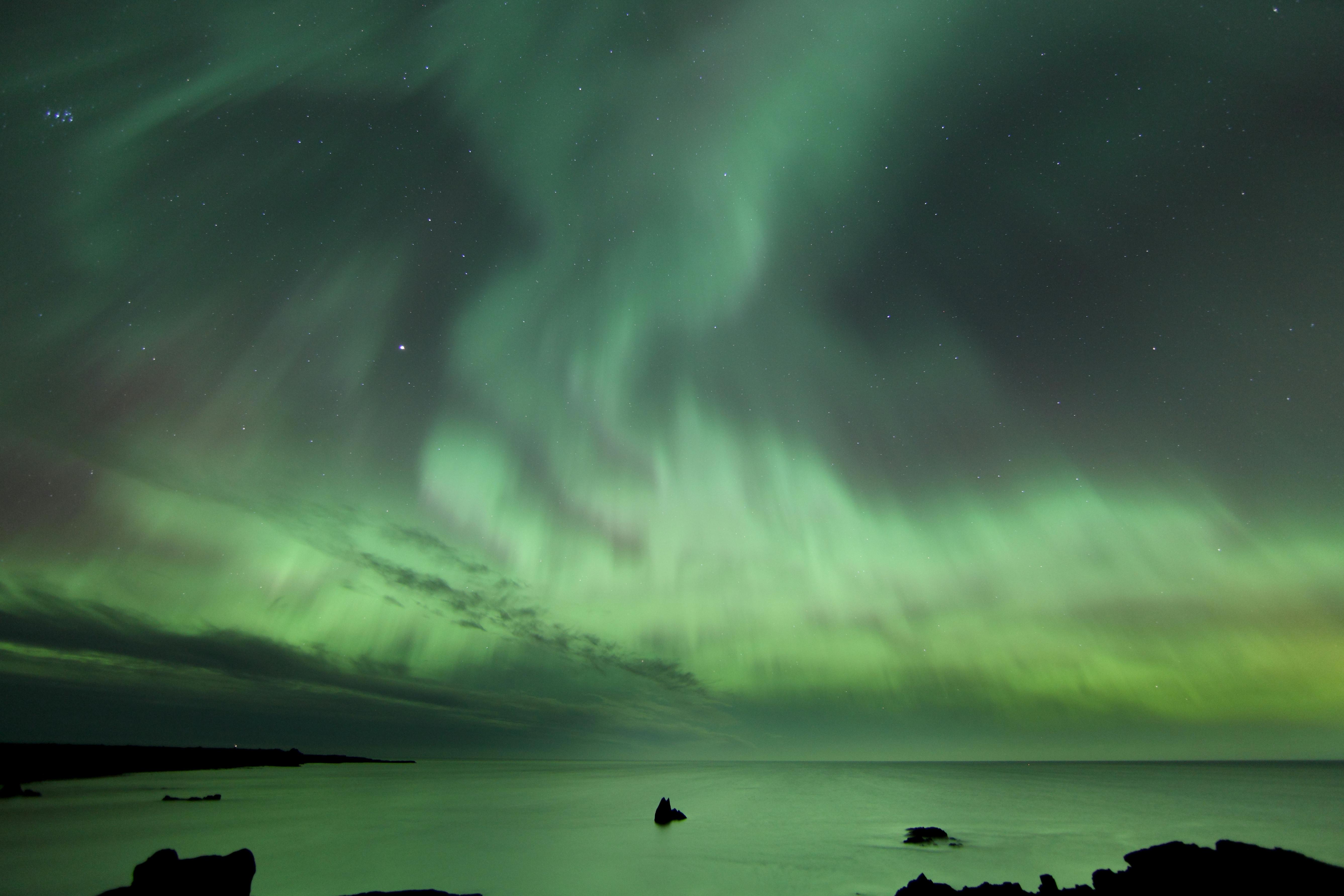 Cielo notturno con aurora boreale verde su un oceano calmo, deboli stelle visibili sullo sfondo.