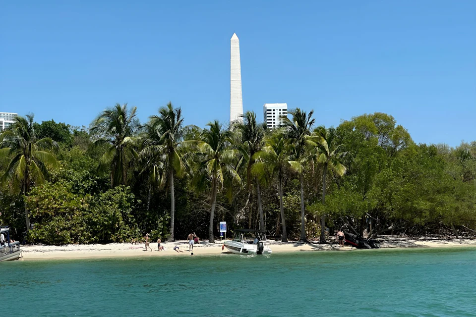 Beach with palm trees, a boat, people on the shore, and a tall white obelisk in the background under a clear blue sky.