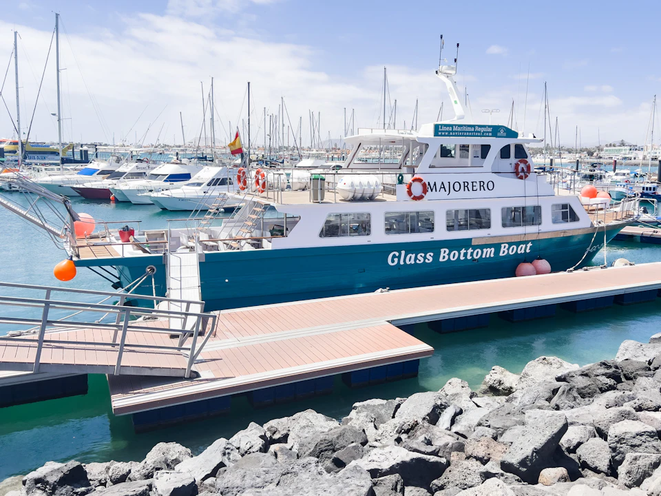 A turquoise and white glass-bottom boat docked at a marina with other boats on a sunny day. Rocks in the foreground.