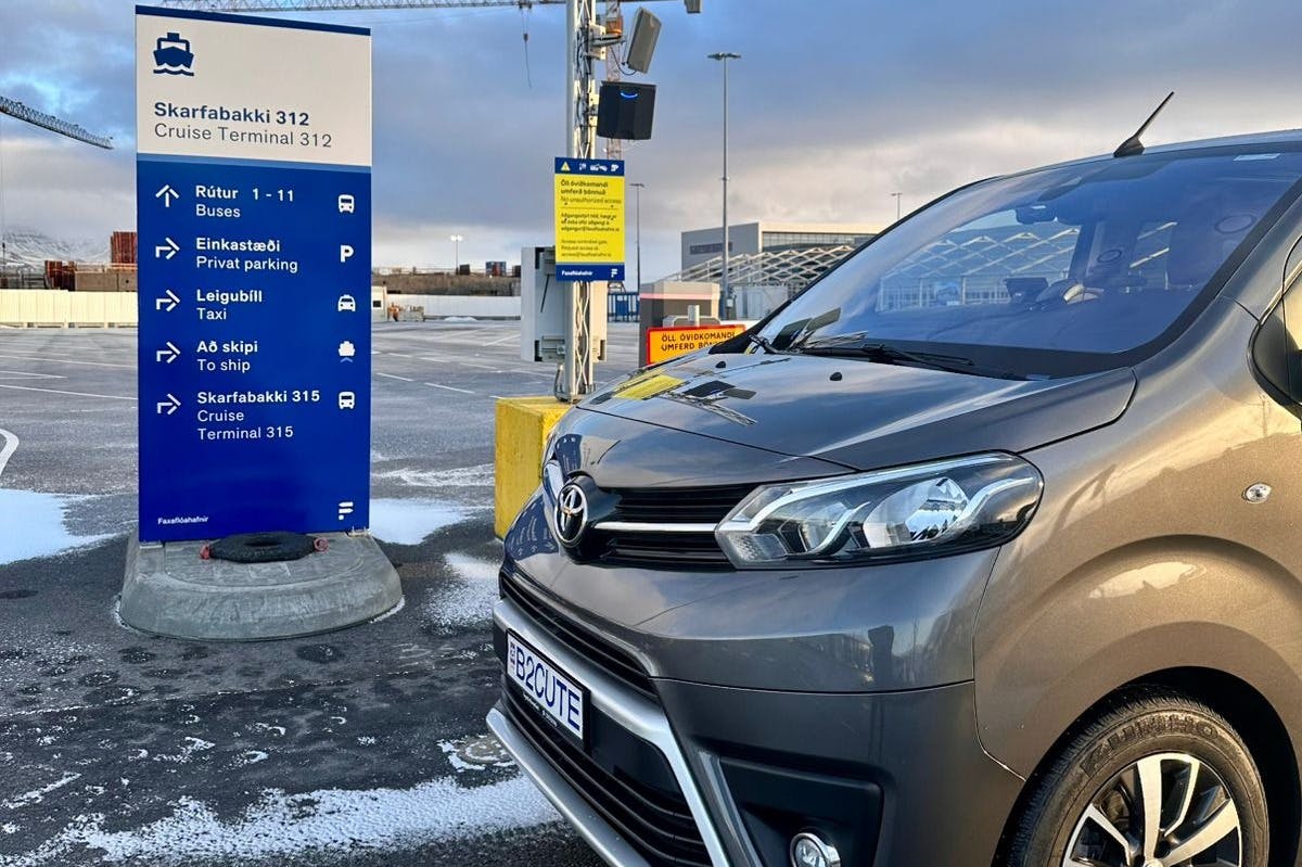 曇り空の下、空港ターミナルの青い案内標識に停まっている「B 8 CUTE」プレートのグレーのトヨタバン。