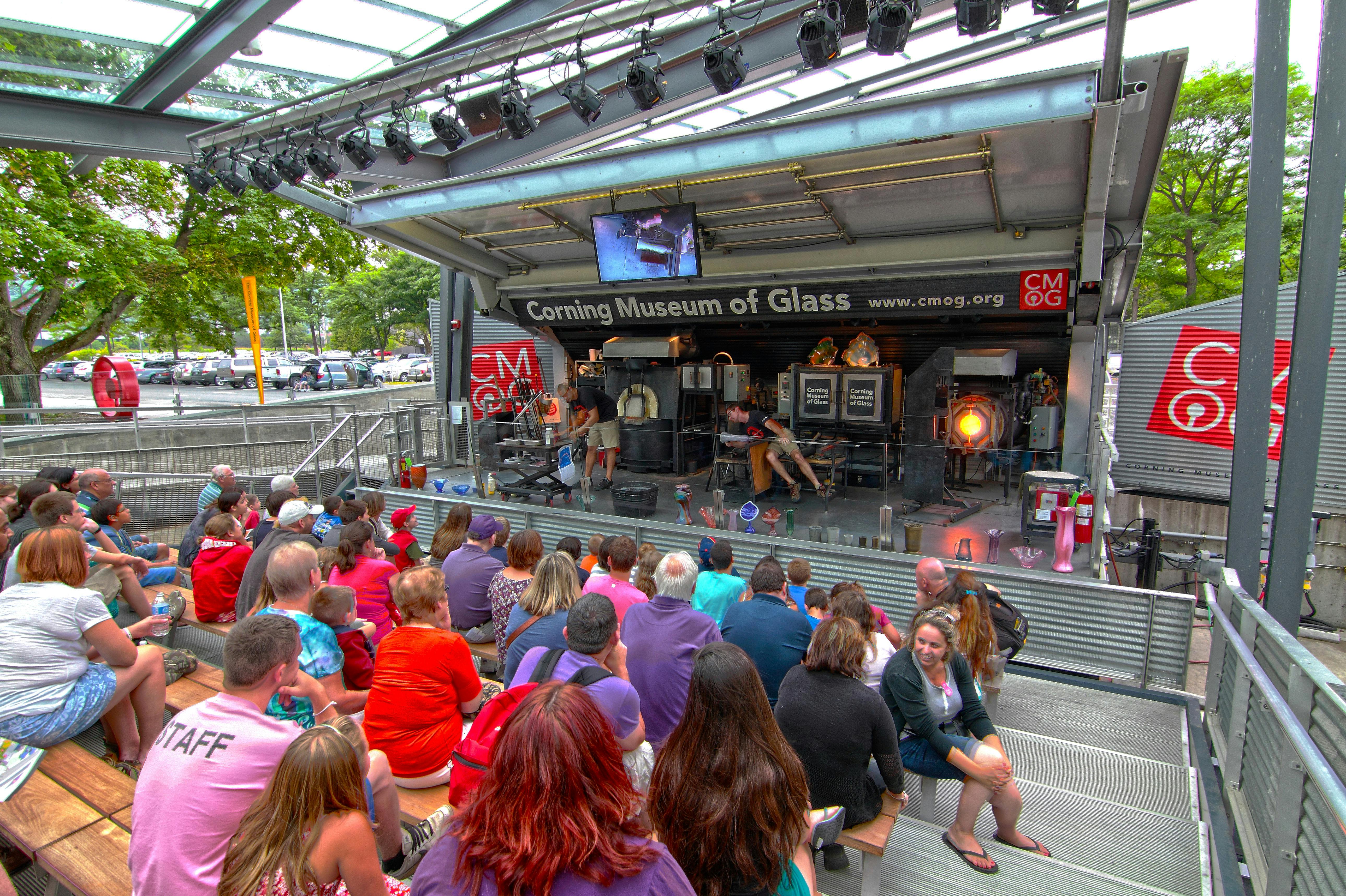 People watching a live glassblowing demonstration at an outdoor exhibit of the Corning Museum of Glass.