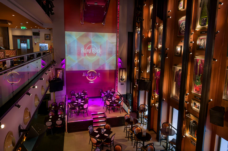 Interior of a Hard Rock Café, featuring a stage, tables with chairs, rock memorabilia on the walls, and colorful lighting.