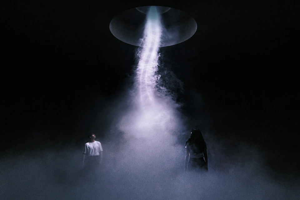 Two people stand in a misty, dark room, looking up at a beam of light descending from a circular opening above.