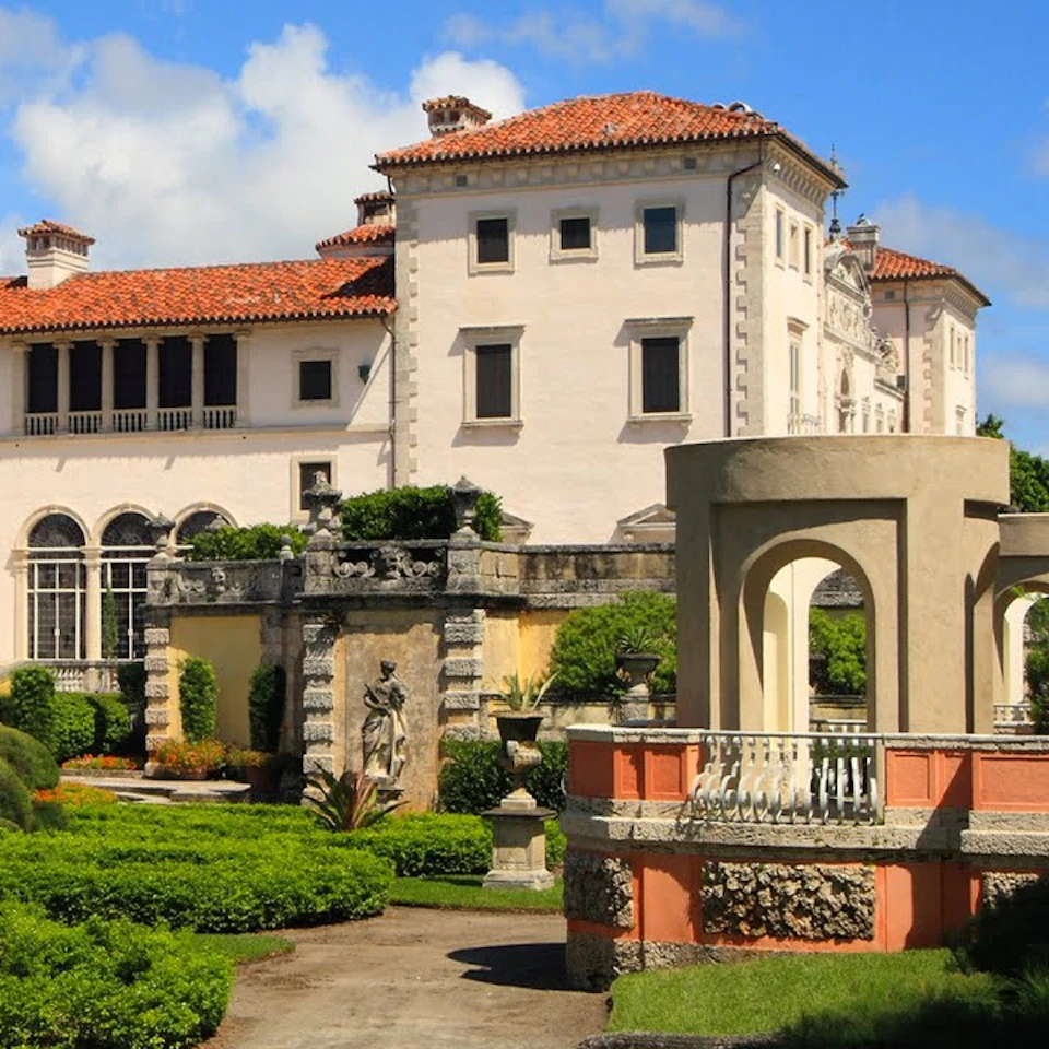 A historic mansion with red-tiled roofs, surrounded by a formal garden with statues, greenery, and archways on a clear day.