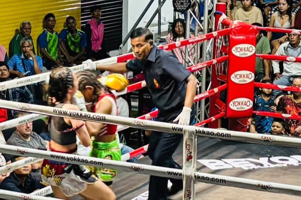 Two fighters compete in a boxing ring surrounded by spectators and judges in an indoor arena.