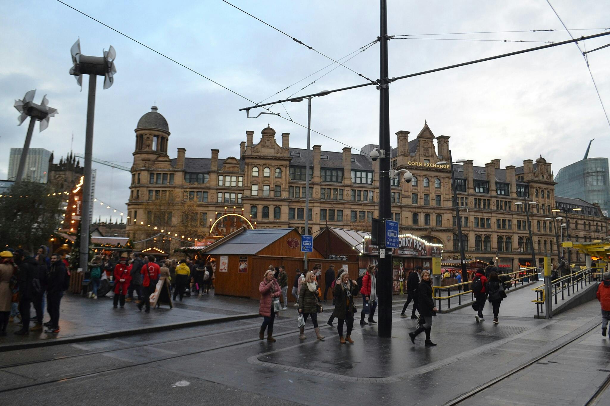 역사적인 건물을 배경으로 번화한 야외 시장의 노점 근처를 걷는 사람들.