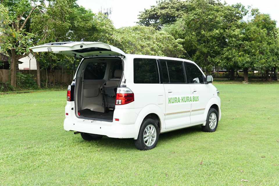 A white van labeled "Kura-Kura Bus" parked on a grassy field with its rear door open, surrounded by trees.