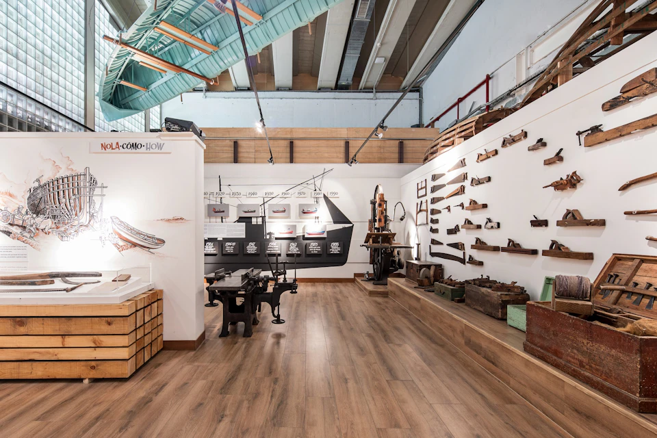 Museum display of vintage woodworking tools on walls and tables in a well-lit room with wooden floors and a high, open ceiling.