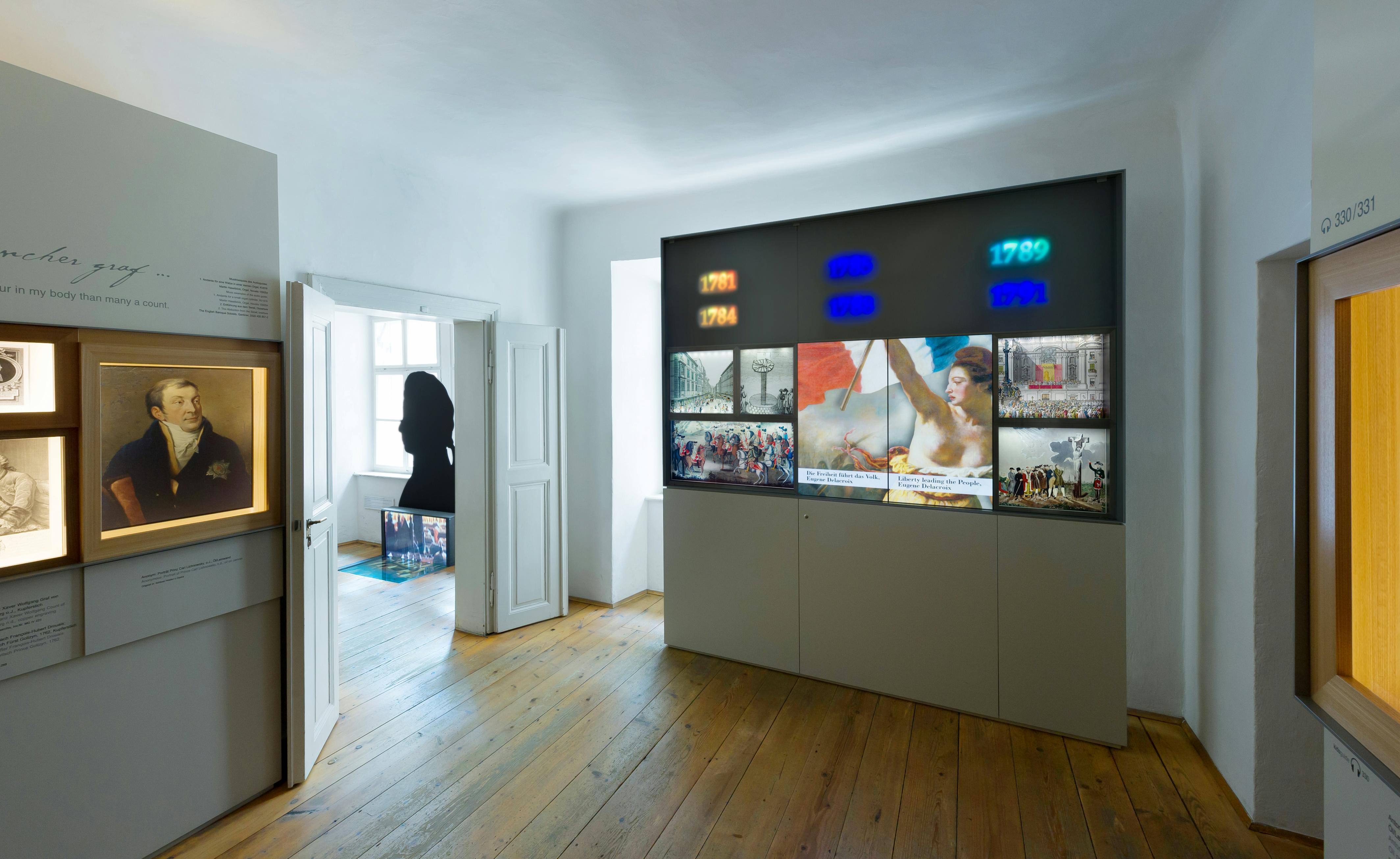 Muzejní místnost s zarámovanými portréty na zdi, otevřenými dveřmi vedoucími do další místnosti a osvětlenými fotografiemi a textem na panelu.