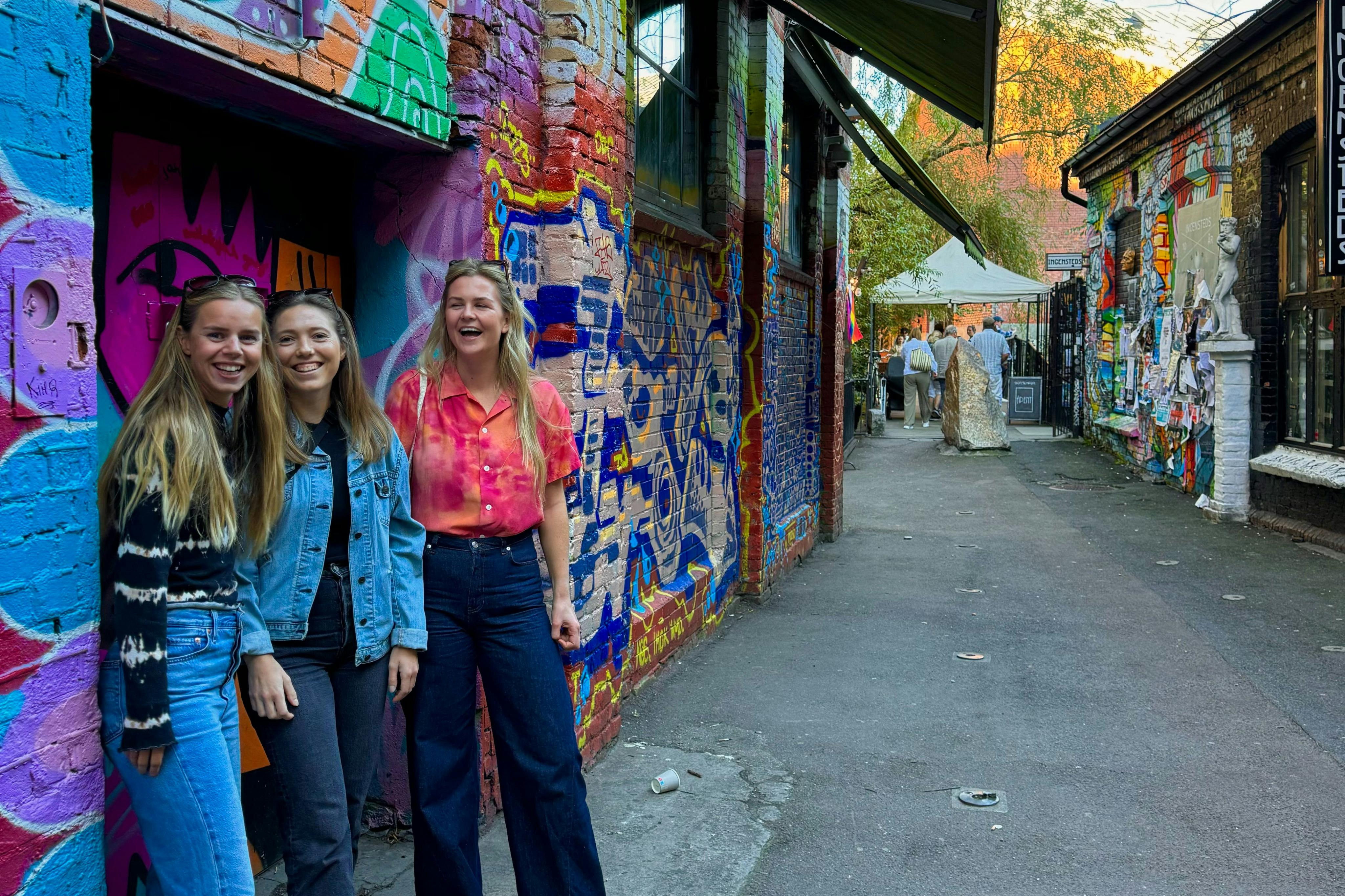 Three women standing together, smiling, in front of a colorful, graffiti-covered wall in an alleyway.