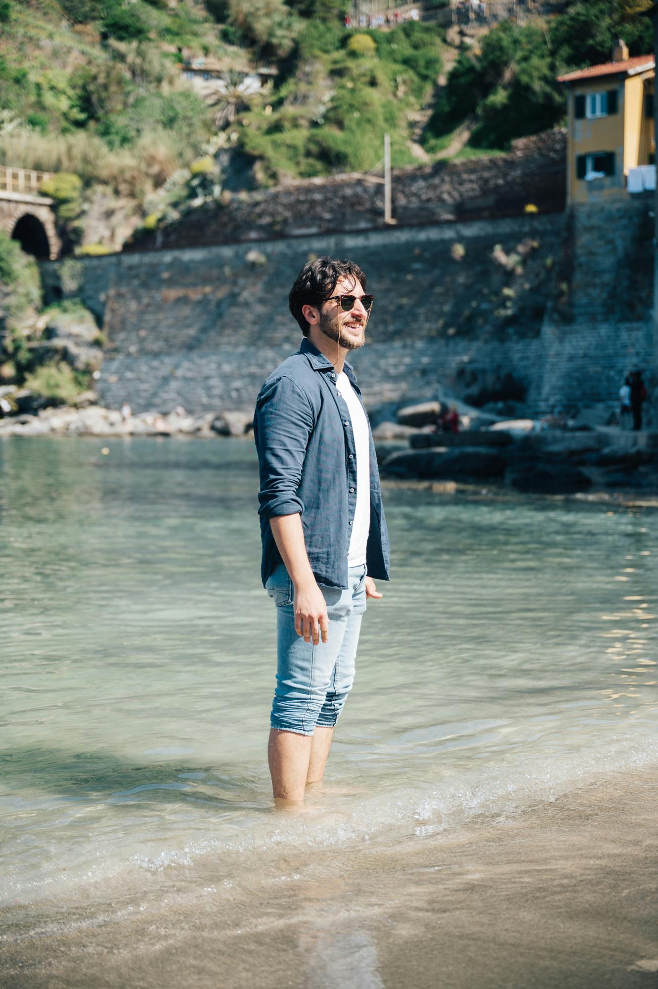 一名男子站在靠近石墙和黄色建筑物的浅水中，戴着太阳镜，穿着休闲服装，背景是绿色植物。