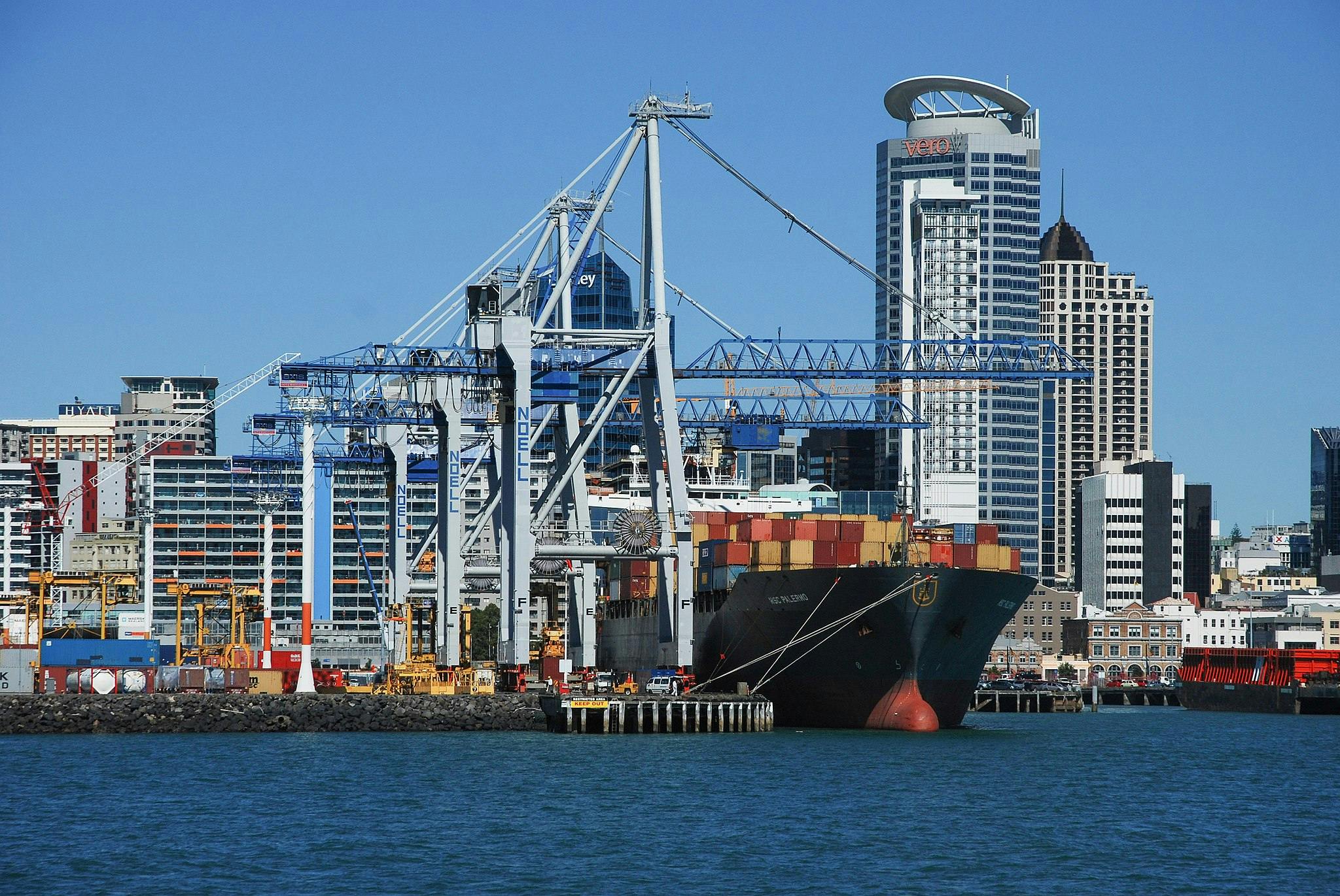 Shipping containers and cranes at a busy port with modern city skyscrapers in the background under a clear blue sky.