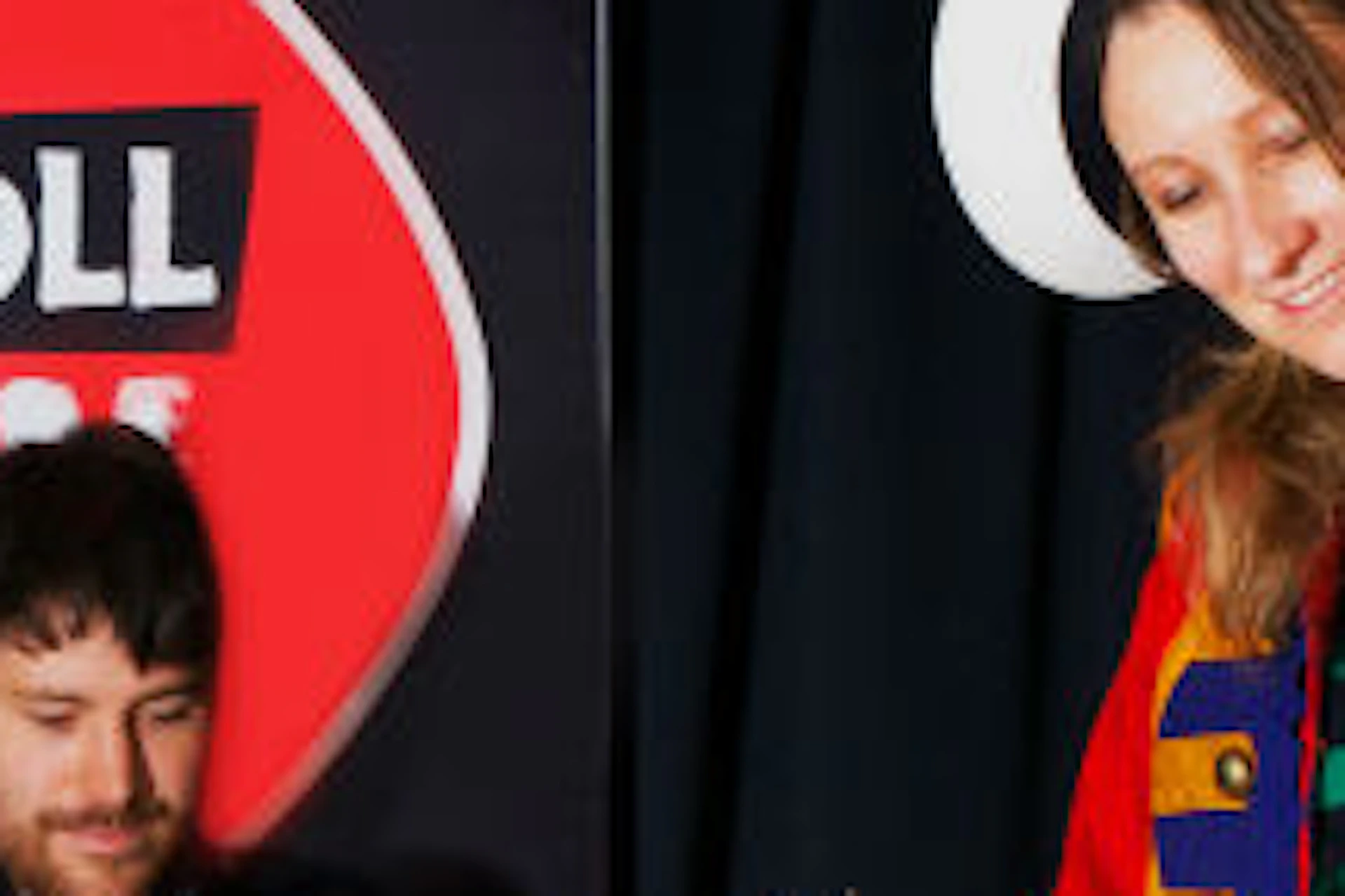 A person in colorful, band-style attire plays a white bass guitar next to a drummer, with an "Irish Rock 'N' Roll Museum Experience" banner behind.
