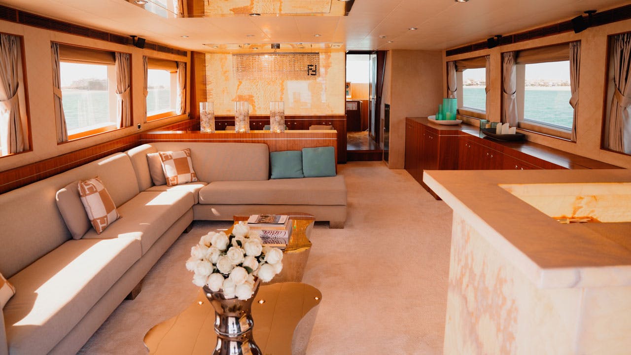 Luxusní obytný prostor na lodi s béžovou sekční pohovkou, skleněným konferenčním stolkem s bílými květy a velkými okny s výhledem na vodu.