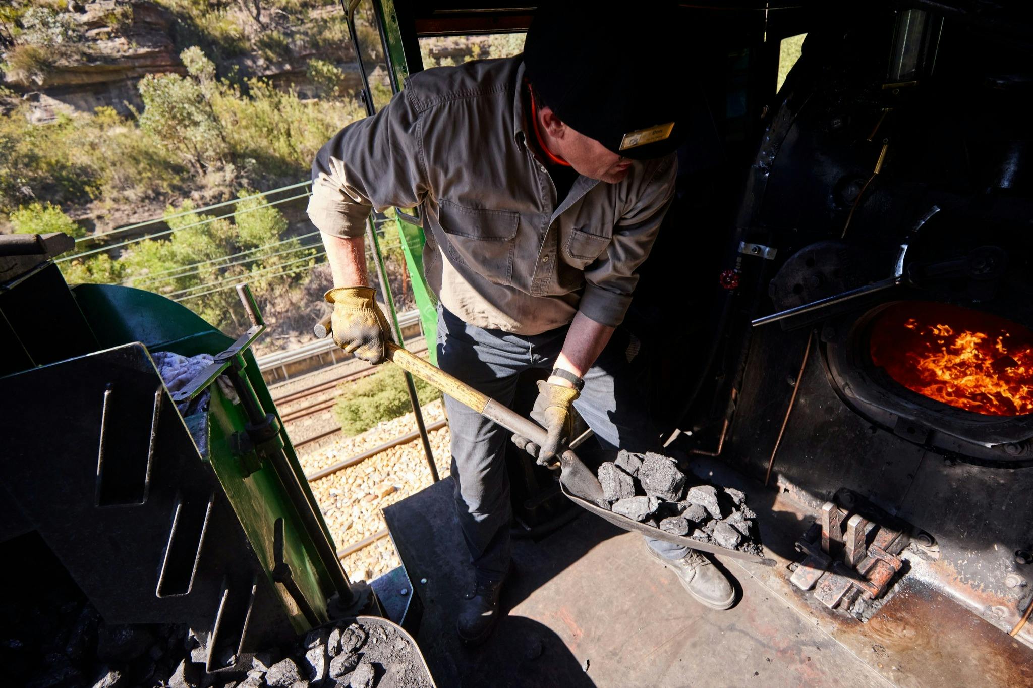 A person shoveling coal into a furnace in a train engine, wearing work gloves and a hat, with railway tracks visible outside.