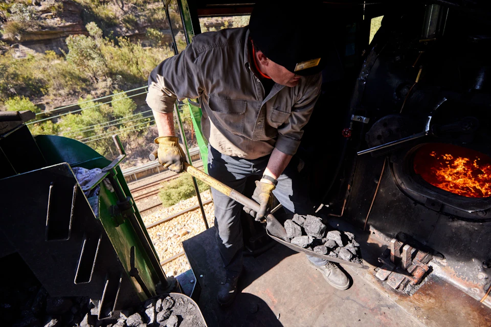 A person shoveling coal into a furnace in a train engine, wearing work gloves and a hat, with railway tracks visible outside.