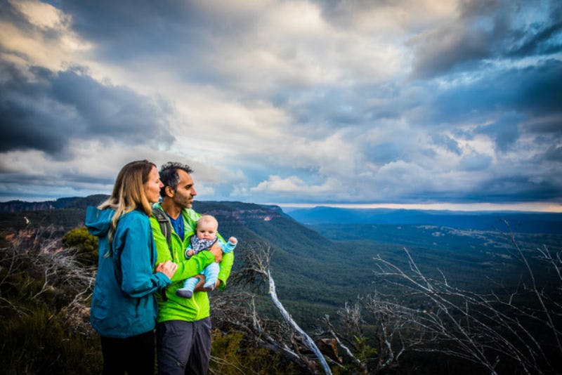 A couple holding a baby stands on a mountain cliff under a cloudy sky, overlooking a vast landscape with green hills.