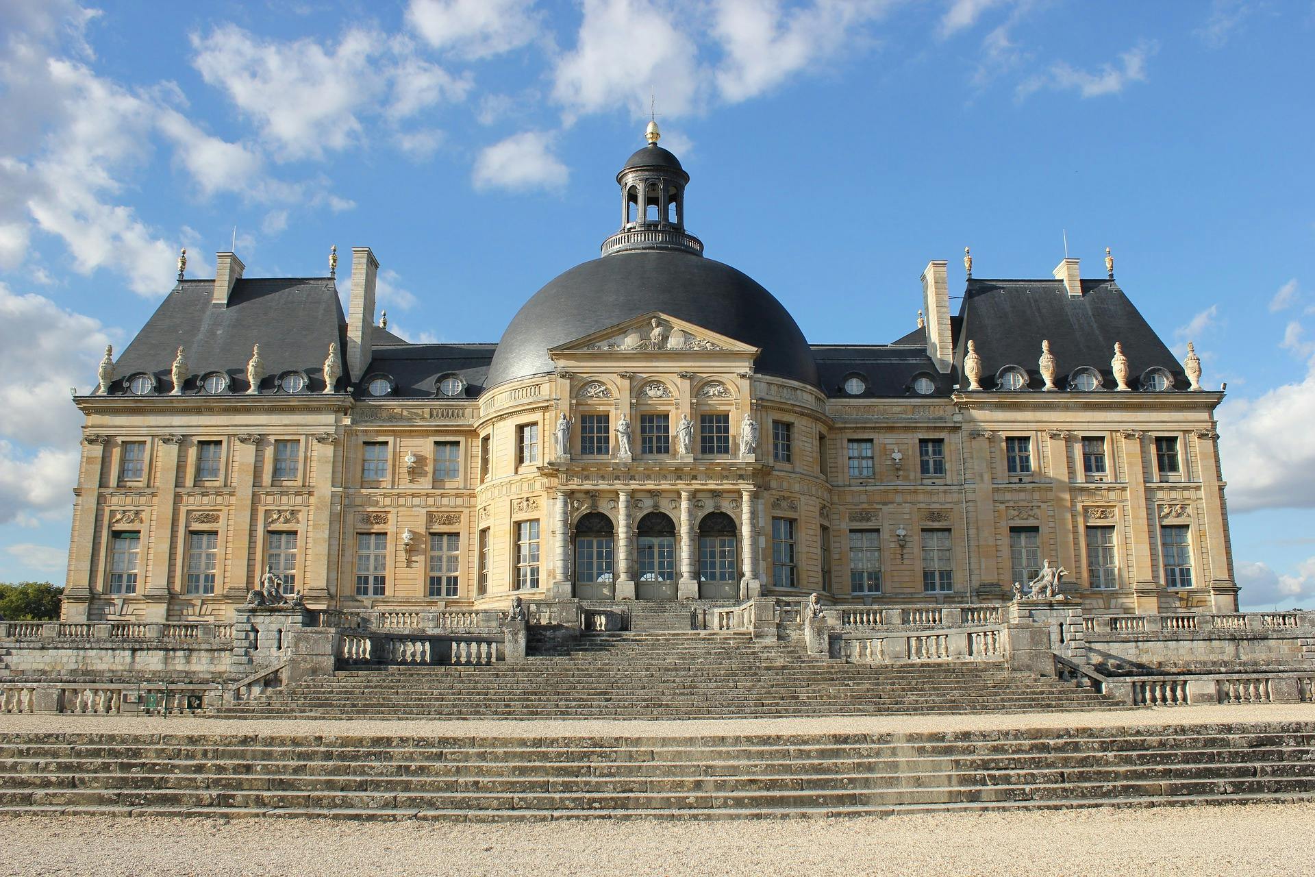 A grand French château with a large dome, ornate architecture, and stone steps leading to the entrance under a blue sky.