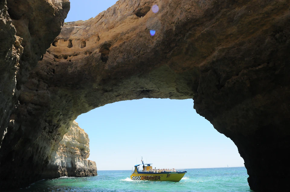 A yellow boat labeled "DREAMER" sails under a large seaside rock arch with a clear blue sky in the background.