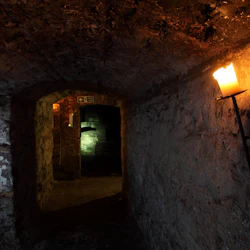 Svagt upplyst stengång med välvt tak och väggar; ett ljus är monterat på höger sida. En upplyst utgångsskylt syns på avstånd.