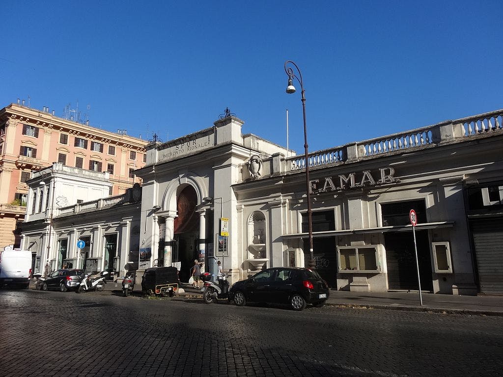 Street scene with scooters, parked cars, and a market building with "SPQR Mercato Rionale" and "FAMAR" signs under a clear blue sky.