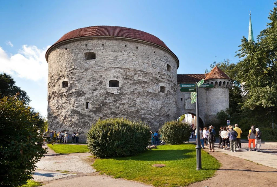 A large, round stone tower with a red-tiled roof and several small windows; people and bushes are in the foreground.