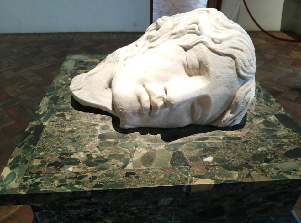 A large marble sculpture of a head resting on its side atop a green, marbled pedestal in a gallery setting.