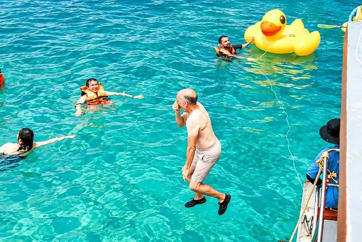 A man jumps into clear blue water near people in life vests and a large inflatable yellow duck.