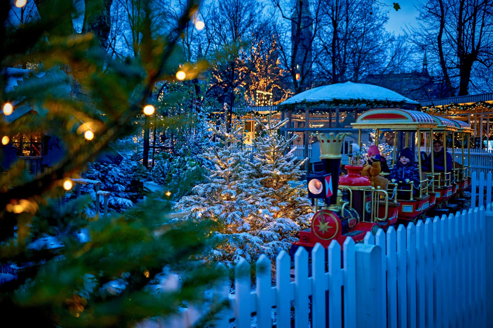 A small train with children rides through a festive, snow-covered park with decorated trees and lights, behind a white fence.