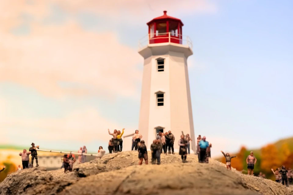 A miniature scene of a lighthouse with a red top, surrounded by small figurines of people on rocky terrain under a blue sky.
