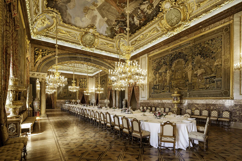 A grand dining hall with ornate chandeliers, a long banquet table, and richly decorated walls and ceiling.
