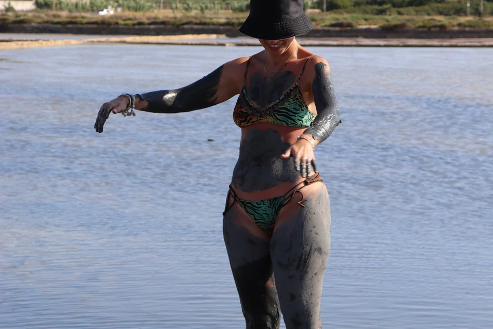 A person in a bikini and hat covered in mud is standing near a body of water, with a natural landscape in the distance.
