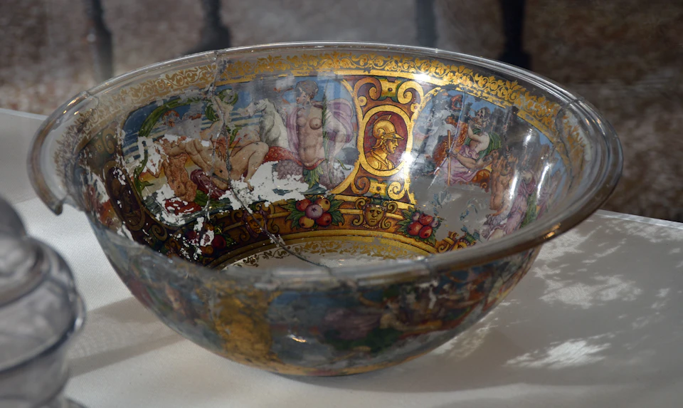 A glass bowl featuring faded, detailed illustrations of classical figures and ornate golden patterns, showing signs of wear.