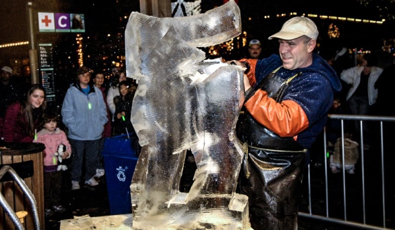 一位戴帽围裙的男士晚上在户外雕刻一座冰雕，背景是人群和节日的灯光。